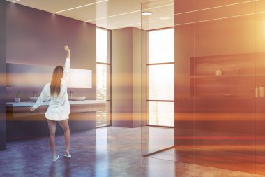 Gri duvarları, beton zemini, duş kabini ve çift lavabosu olan modern banyoda duran genç bir kadın. Tonu ayarlanmış resim