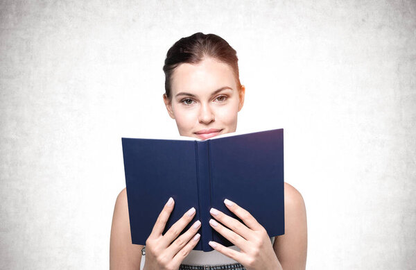 Portrait of young businesswoman standing with blue book over concrete wall background. Concept of knowledge