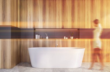 Modern banyoda tahta ve gri duvarları, beton zemini ve rahat küveti olan bulanık genç bir kadın yürüyor. Tonu ayarlanmış resim