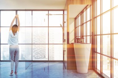 Modern banyoda tahta ve gri duvarları, beton zemini, küveti ve lavabosu olan genç bir kadın. Tonu ayarlanmış resim