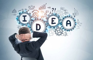 Gri duvarın yanında oturan rahat genç iş adamının arkası. Üzerinde yaratıcı iş fikirleri ve dişliler çizilmiş. Beyin fırtınası kavramı