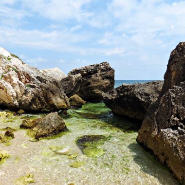 Kayalar, berrak deniz ve beyaz bulutlar ile mavi gökyüzü