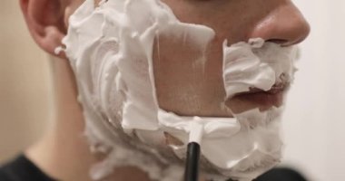 Adam köpükle tıraş oluyor. Banyoda tek kullanımlık tıraş bıçağı kullanıyor. Yakın plan, yan görüş..