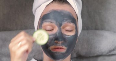 Yüzünde gri maske olan genç kız gözlerine salatalık dilimleri sürüyor..