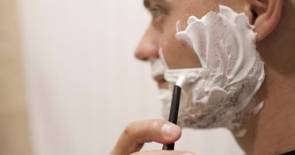 Молодой человек бреется с помощью одноразовой бритвы в ванной комнате, крупный план, вид сбоку. — стоковое видео