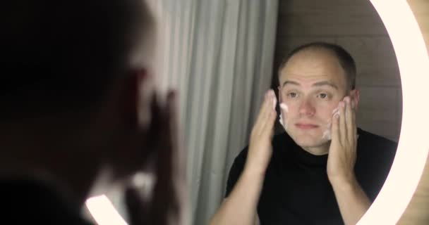 Manusia menerapkan busa pembersih di wajah untuk mencucinya dan melihat ke cermin. — Stok Video