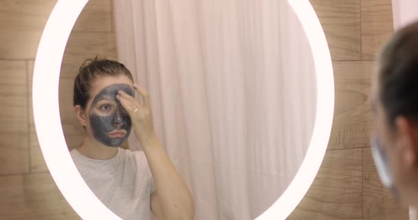 Γυναίκα εφαρμόζει πηλό ενυδατική μάσκα στο πρόσωπο στο μπάνιο κοιτάζοντας καθρέφτη. — Αρχείο Βίντεο