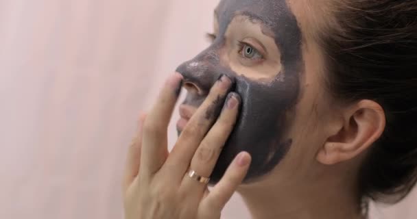 Jovem mulher está aplicando máscara cinza argila facial no rosto, close-up vista lateral . — Vídeo de Stock