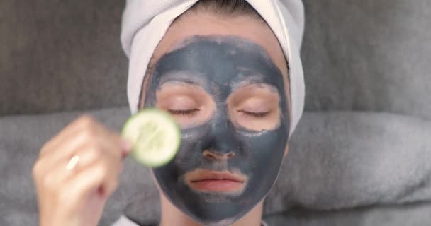 Młoda dziewczyna z szarą maską na twarzy kładzie plastry ogórka na oczy, spa. — Wideo stockowe
