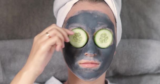 Młoda kobieta z glinianą maską na twarzy biorąca ogórek z oka i jedząca go. — Wideo stockowe