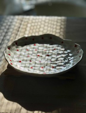 El yapımı, kırmızı kalp desenli bir seramik tabak. Güneş ışığı ve örülmüş masa örtüsünün üzerindeki gölgelerle güzelce vurgulanmış..
