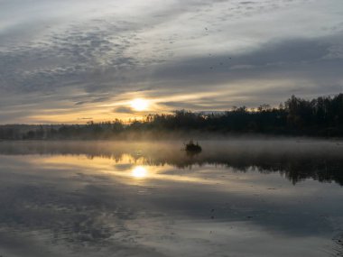 Sonbahar sabahının erken saatlerinde, su ve zemin üzerindeki beyaz sis, arka planda güzel renkli ve bulanık ağaç siluetleri