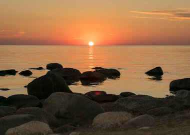 Deniz kenarında turuncu gün batımı, deniz zemininde siyah taş siluetler, yaz akşamı