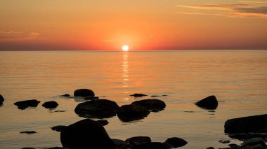 Deniz kenarında turuncu gün batımı, deniz zemininde siyah taş siluetler, yaz akşamı