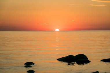 Deniz kenarında turuncu gün batımı, deniz zemininde siyah taş siluetler, yaz akşamı