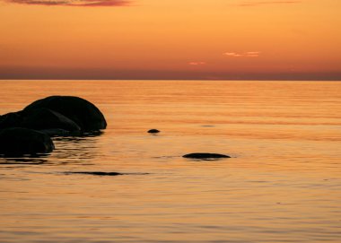 Deniz kenarında turuncu gün batımı, deniz zemininde siyah taş siluetler, yaz akşamı