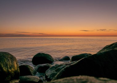 Deniz kenarında turuncu gün batımı, deniz zemininde siyah taş siluetler, yaz akşamı