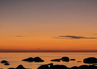 Deniz kenarında turuncu gün batımı, deniz zemininde siyah taş siluetler, yaz akşamı