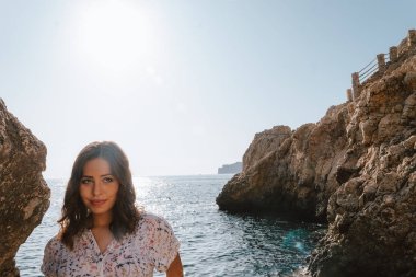 Maceracı genç latin kadın deniz kayalar üzerinde yan bakıyor. Uçurumdaki kadın, gün batımını olan. Turkuaz mavisu ile Akdeniz'de tatilde denize yakın Jovial.