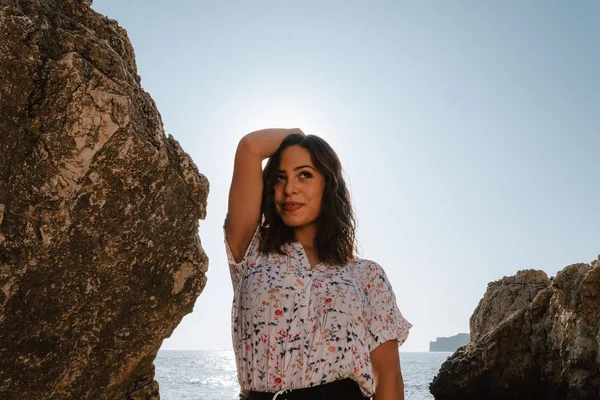 Adventurous young latin woman grooming her hair looking straight ahead smiling. Woman on cliff, with the sunset. Jovial near the sea on vacation in the Mediterranean with turquoise blue water. — Stock Photo, Image