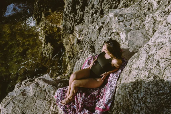 Jovem hipster deitada sobre as rochas em um maiô sobre toalha de praia. Jovem deitada perto do mar com óculos de sol vintage . — Fotografia de Stock