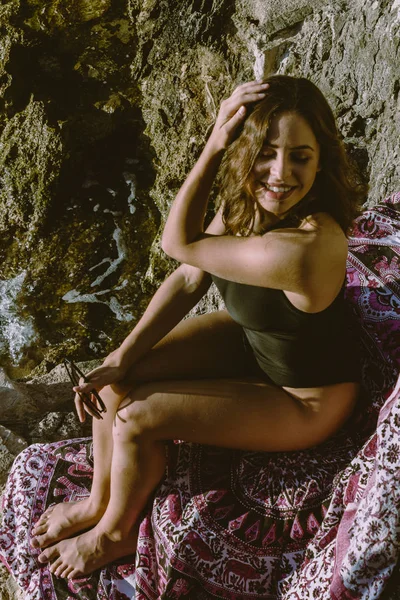 Genç hipster kadın plaj havlusu üzerinde bir mayo içinde kayalar üzerinde yatıyordu. Genç kadın vintage güneş gözlüğü ile deniz kenarında yatan. — Stok fotoğraf