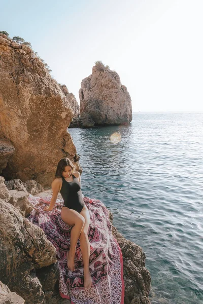 Genç hipster kadın plaj havlusu üzerinde bir mayo içinde kayalar üzerinde yatıyordu. Genç kadın vintage güneş gözlüğü ile deniz kenarında yatan.