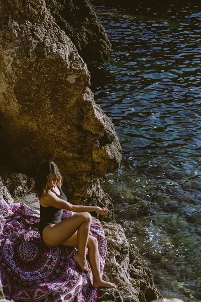 Jovem hipster deitada sobre as rochas em um maiô sobre toalha de praia. Jovem deitada perto do mar com óculos de sol vintage . — Fotografia de Stock