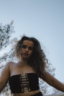 Siyah bluzlu, kıvırcık saçlı, arka planda ağaçlı bir kadın..