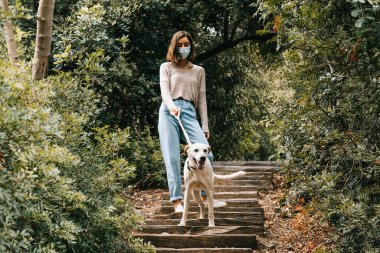Sokakta yüz maskesi takan krem rengi bir köpeği gezdiren genç bir kadın. Yumuşak renkler. Yeni normal açık hava kavramı.