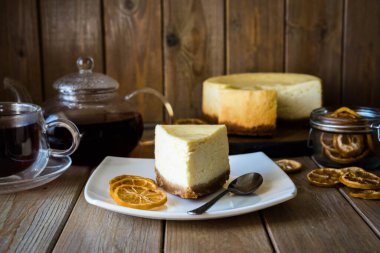 Limon dilimli beyaz tabakta bir dilim limonlu cheesecake, bir fincan sıcak çay ve ahşap masada çay ile bir bardak çaydanlık.