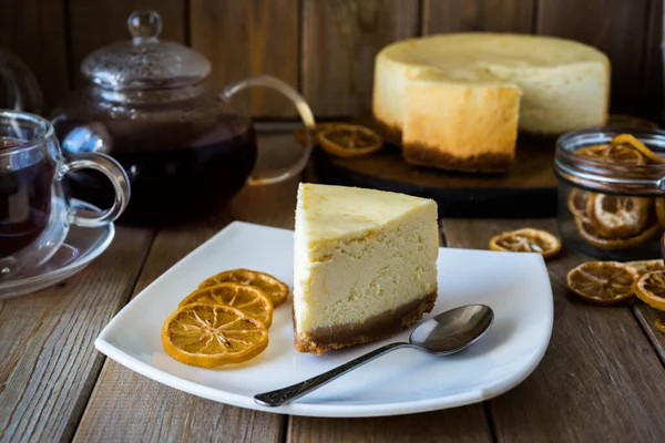 Limon dilimleri ve kaşıkla beyaz tabakta bir dilim cheesecake. Arka planda sıcak çay ve bir fincan çayla birlikte cam bir çaydanlık var.