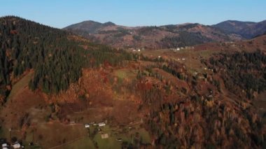 Dağ köyü yeşil yamaçtaki ender evler yoğun sonbahar ormanlarıyla çevrili güneş ışığı gökyüzü manzarası ile aydınlatılıyor. Karpat Dağları, Ukrayna güzellik doğası. Seyahat, sonbahar tatili. 4k