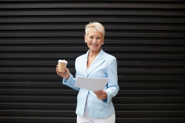 Elinde belgeler ve kahve içen ve dışarıda kameraya gülümseyen başarılı bir iş kadını portresi.