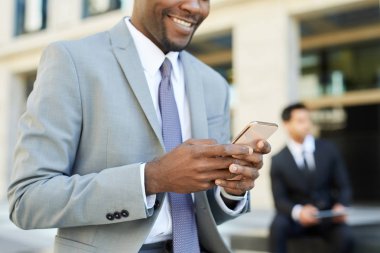 Gülümseyen Afrikalı işadamı arka planda meslektaşıyla otururken cep telefonu kullanıyor ve kahve içiyor.