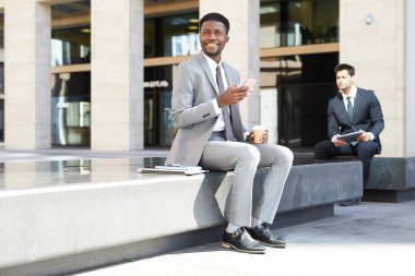 Gülümseyen Afrikalı işadamı arka planda meslektaşıyla otururken cep telefonu kullanıyor ve kahve içiyor.