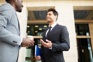 Takım elbiseli iki genç iş adamı şehirde yürürken konuşuyor.