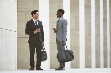 Takım elbiseli iki çok ırklı işadamı ellerinde poşetlerle şehirde dikilirken kahve içip birbirleriyle konuşuyorlardı.