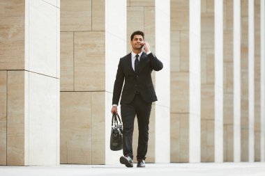 Siyah takım elbiseli genç bir işadamı şehirde yürürken cep telefonuyla konuşuyor. 