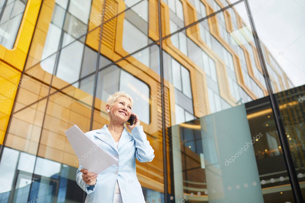 Successful mature businesswoman with short hair holding documents and talking on mobile phone in the city with modern office building in the background