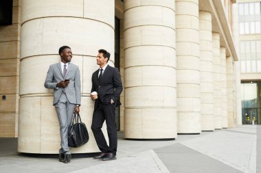 Takım elbiseli iki genç iş adamı modern binanın yanında dikilirken konuşuyor. 