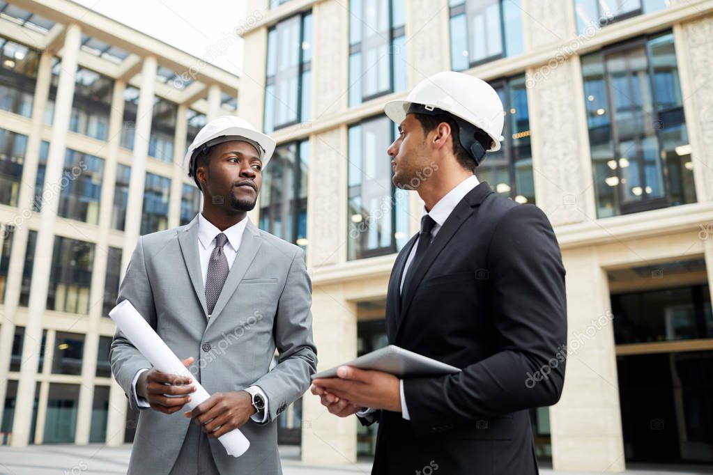 Two multiethnic engineers in hardhats discussing new project and working in team while standing in big city among modern buildings 