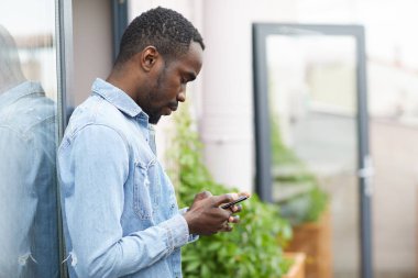 Mavi tişörtlü, ciddi bir Afrikalı iş adamı ofiste dururken internette cep telefonuyla çalışıyor.