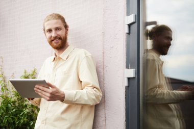 Dijital tablet kullanan, dışarıda durup binanın duvarına yaslanırken kameraya gülümseyen kızıl saçlı genç adamın portresi.
