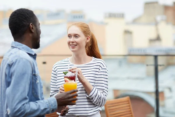 African cheerful man drinking fruit juice and talking to young woman standing outdoors