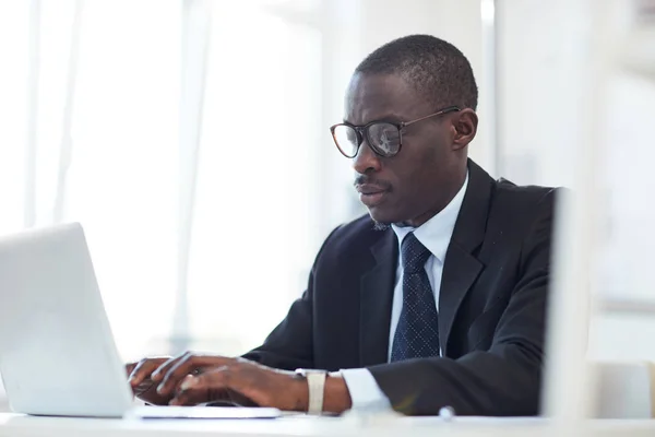 テーブルに座って昼にオフィスのノートパソコンに入力する眼鏡の深刻なアフリカのビジネスマン — ストック写真