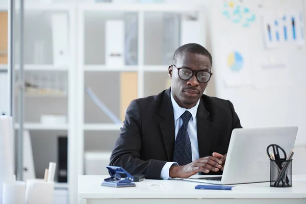 眼鏡や黒いスーツを着たアフリカ人ビジネスマンがノートパソコンの前の職場に座り オフィスでカメラを見ている姿 — ストック写真