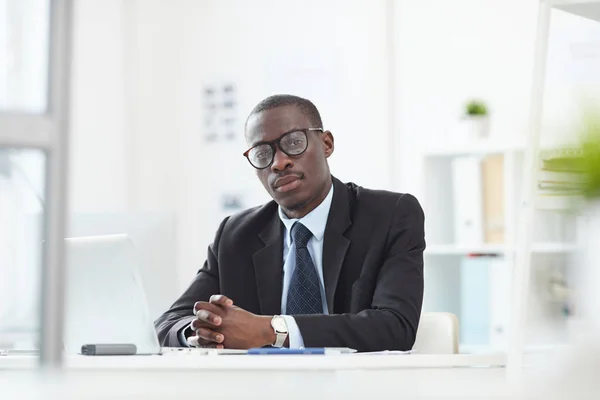 Portrait of African serious manager in eyeglasses sitting at table with laptop and looking at camera at office