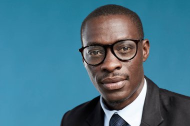 Gözlüklü ve takım elbiseli genç Afrikalı işadamının portresi mavi arka planda kameraya bakıyor.