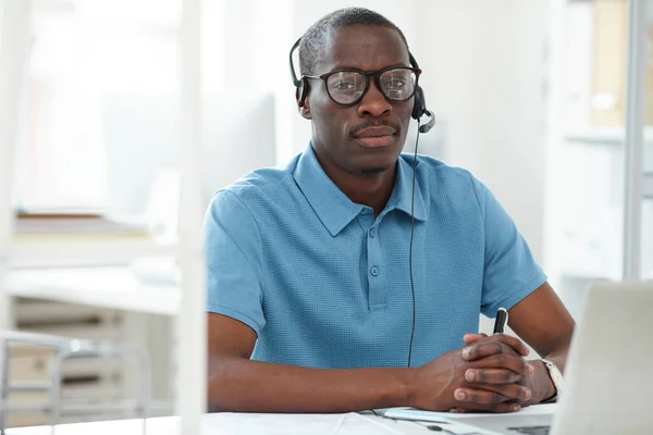 ノートパソコンの前の職場に座ってオフィスでカメラを見ているヘッドフォンや眼鏡をかけた若いアフリカ人デザイナーの肖像画 — ストック写真
